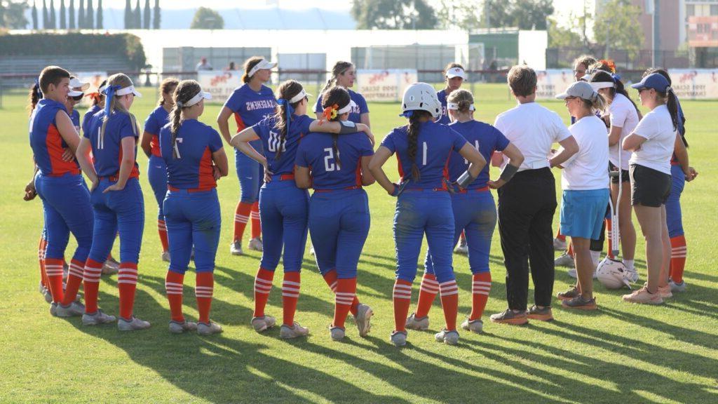 The women's softball team stand in a circle with their coaches on a field. The team wears blue Sagehens uniforms with orange and white socks.