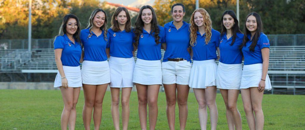 Six members women's golf stand in a row on a field. They wear blue shirts and white shorts and skirts for their uniform.